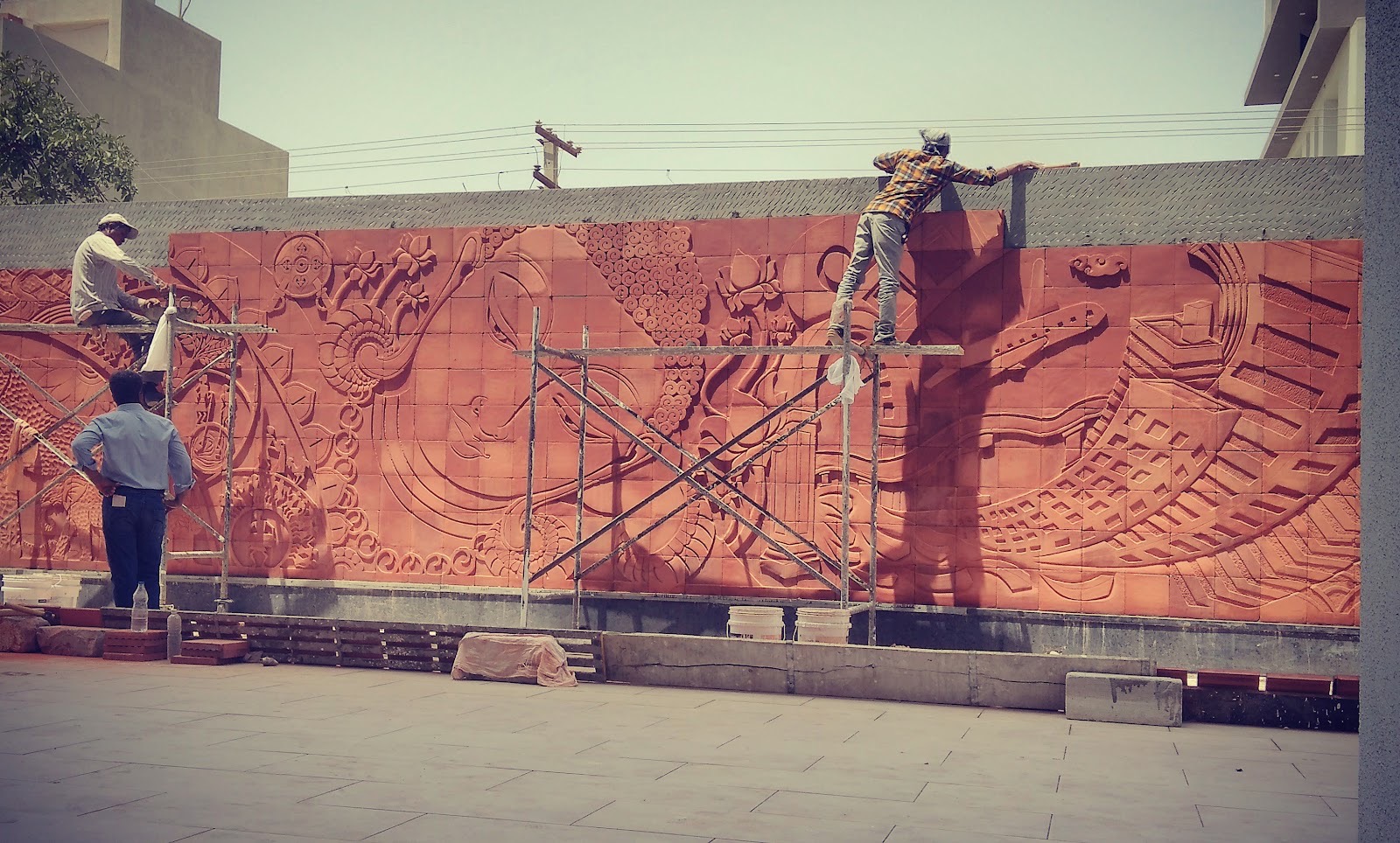 Red relief mural wall of swirling motifs being installed by artisans on scaffolding